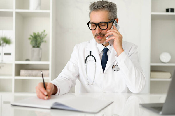 The mature doctor is pictured in a moment of ease, smiling while speaking on the phone, a stethoscope draped around his neck, portraying a friendly and reassuring medical consultation, taking notes