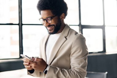 Genç işadamı telefonuna bakarken gülümsüyor, başarılı bir girişimcinin bağlı kalmasının bir sembolü, parlak, havadar bir ofisin arka planı. Erkek ofis çalışanı kapalı alanda telefon kullanıyor.
