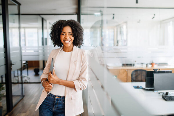 A confident young woman with a bright smile stands by a glass partition, holding the laptop in her hand, embodying professionalism and readiness. The concept of career advancement