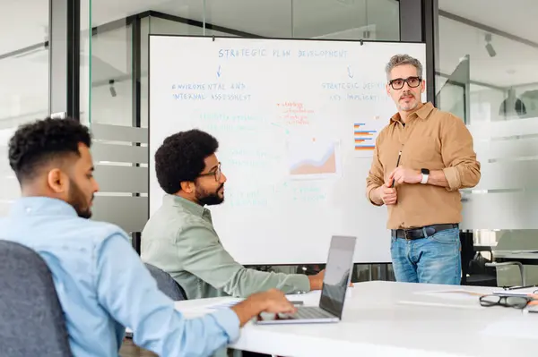 Mature man in a casual shirt confidently presents a strategic plan on a whiteboard to his attentive colleagues in bright, modern office, exemplifying focused team collaboration and strategic planning