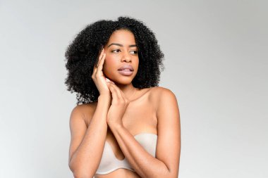 Düşünceli bir Afro-Amerikalı kadın yanağına dokunuyor, sakin bir ifadeyle, pürüzsüz bir arkaplana bakıyor. Fotoğraf, sessiz bir tefekkür ve nazik bir iç gözlem hissi veriyor..