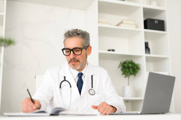 Focused grey-haired doctor taking notes while using laptop, reviewing patient files or medical literature. His concentration reflects diligence and expertise expected in modern medical care