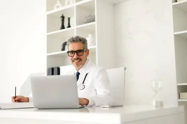 Mature Doctor Friendly Demeanor Types Laptop While Taking Notes Showcasing Stock Photo