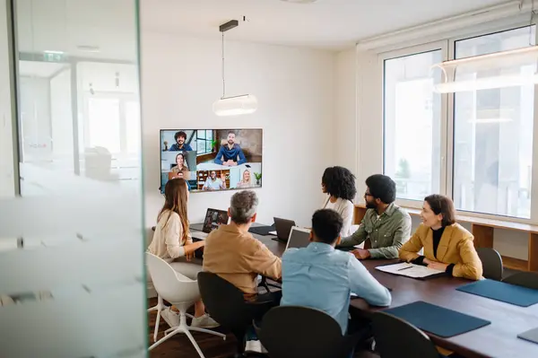 Stock image A team actively engages with remote colleagues via a large screen, illustrating the modern hybrid work model where technology bridges the gap between in-office and remote team members