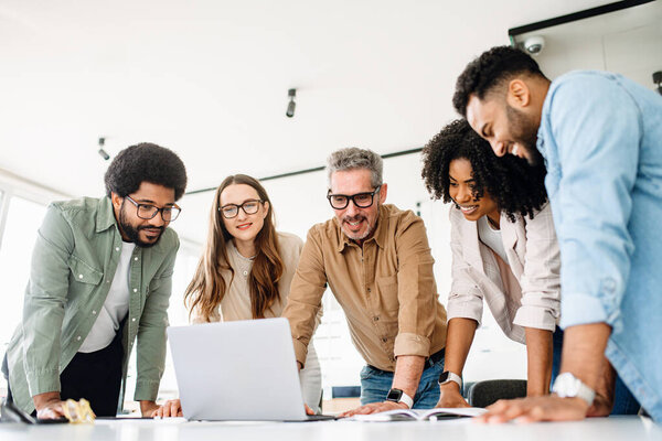 A group of diverse professionals is standing around a laptop, highlighting a moment of team collaboration and digital strategy planning, solving project tasks, or analyzing the result