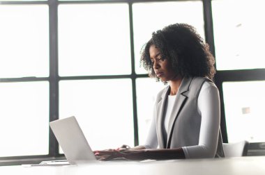 Afro-Amerikalı bir kadın oturur ve dizüstü bilgisayar kullanır, silueti, arkasındaki geniş pencerelerin geometrik çizgileriyle çerçevelenmiştir, modern çalışma alanlarının temaları ve odaklanmış profesyoneller için mükemmeldir..