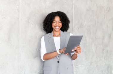 Profesyonel kıyafetli coşkulu bir Afro-Amerikalı iş kadını dijital tablet taşıyor. Işıl ışıl gülüşü ve göz teması, kurumsal ortamlarda yaklaşılabilirlik ve teknoloji meraklılığı olduğunu gösteriyor.