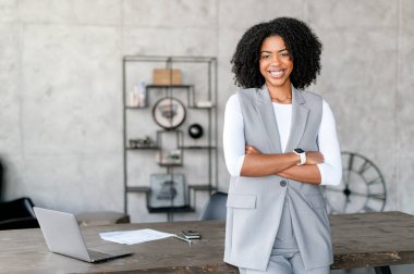 Işıldayan bir Afro-Amerikalı kadın, kollarını, modern profesyonellik ve yaklaşılabilirliğin bir karışımını temsil eden, şık bir iş-gündelik yeleği ve beyaz uzun kollu gömleğiyle çapraz duruyor.