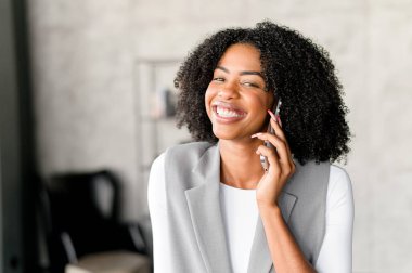 Parlak bir gülümsemeyle, bu Afro-Amerikalı iş kadını telefonuyla neşeli bir sohbete girişiyor, modern ofis ortamında bağlantı ve ulaşılabilirliği yansıtıyor..