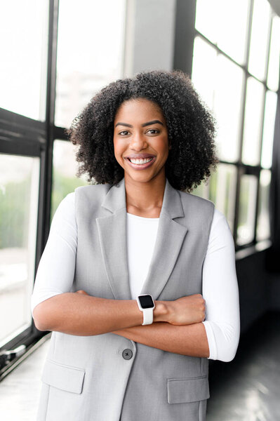 A poised African-American businesswoman with a cheerful demeanor stands arms crossed in a modern office, her smartwatch hinting at a fusion of style and technology in the workplace.