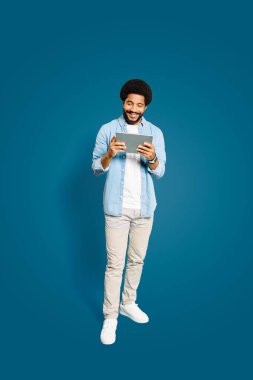 Brezilyalı adam tablet ekranını seyrediyor, gülümsüyor, cihazı rahat bir şekilde tutuyor, modern teknolojik aletlerin konforlu ve sezgisel kullanımının bir göstergesi olarak, mavi üzerinde tam boy izole edilmiş.