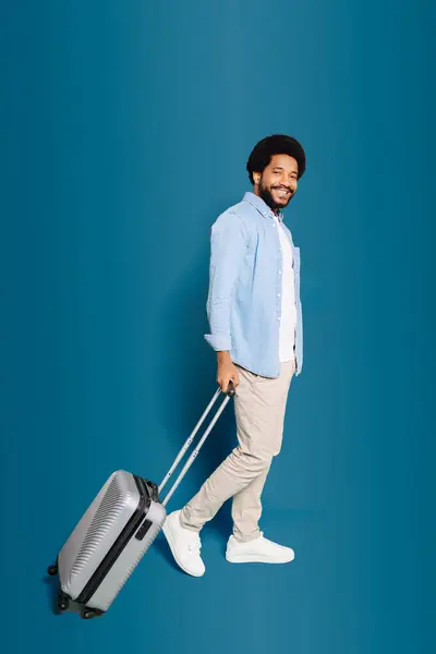 Cheerful man walking with a suitcase in hand, suggests themes of travel, adventure, and the joy of new experiences. The blue backdrop offers a clear space for travel agencies or holiday promotions
