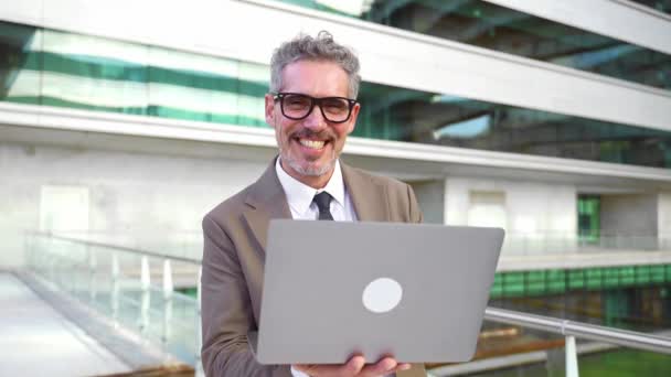 一位穿着棕色西装 自信的60多岁的老商人站在户外 拿着笔记本电脑 带着迷人的微笑看着相机 展示着经验与现代技术的融合 — 图库视频影像