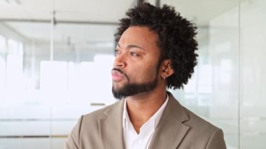 Dostane bir gülümsemesi olan karizmatik bir Afro-Amerikalı işadamı modern bir ofis ortamında durur, profesyonellik ve samimiyet yayar, hayali bir kenara bakar ve sonra gülümseyerek bir kameraya bakar.