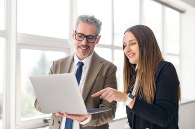 Genç bir bayan profesyonel bilgisayar ekranına işaret ederken, neşeli bir ifadeye sahip kıdemli bir işadamı, ışık dolu bir ofis alanında, işbirlikçi ve eğitici bir anın göstergesidir.