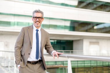 Olgun gri saçlı yakışıklı bir işadamı ofis balkonunda duruyor ve kameraya bakıyor. Çekici gülüşü ve doğrudan bakışları erişilebilirliği ve açıklığı gösteriyor..
