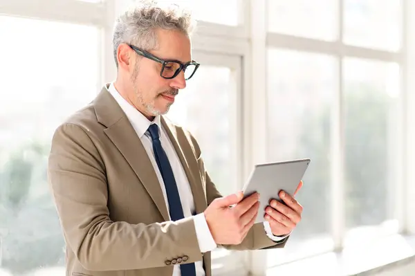 stock image A distinguished businessman in a tailored beige suit confidently operates a tablet, reflecting the dynamic integration of technology in business, highlighting the fusion of experience and innovation