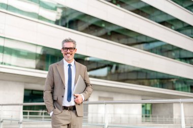 Takım elbiseli kıdemli işadamı dizüstü bilgisayarını rahat bir şekilde kolunun altında tutuyor, dışarıda modern cam bir binanın önünde duruyor, iş dünyasının hareketliliğini somutlaştırıyor.