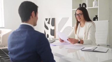 Beyaz ceketli bir kadın, modern, parlak bir ofiste masasının karşısında mavi takım elbiseli bir adamla belgeleri inceliyor. Sahne iş görüşmesi ya da iş görüşmesi öneriyor