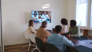 Çeşitli meslektaşlardan oluşan bir grup modern bir konferans odasında toplanarak geniş bir ekranda görüntülenen bir video konferans görüşmesine aktif olarak katılıyor. Gösterilerle tanışmak işbirlikçi bir çalışma ortamı