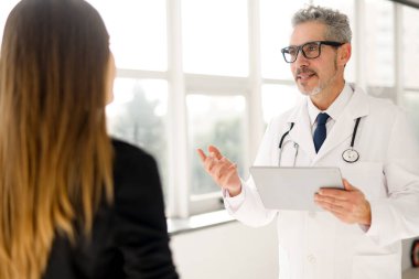 Pencere manzaralı iyi ışıklandırılmış bir ofiste, olgun, gri saçlı bir doktor kollarını kavuşturup, tıp alanında profesyonellik ve deneyimi resmediyor..