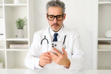 Üst düzey bir doktor akıllı telefonuna odaklanır, hasta bilgilerini gözden geçirir veya koordine eder, mobil teknolojinin modern tıbbi uygulamalara entegrasyonunu gösterir.