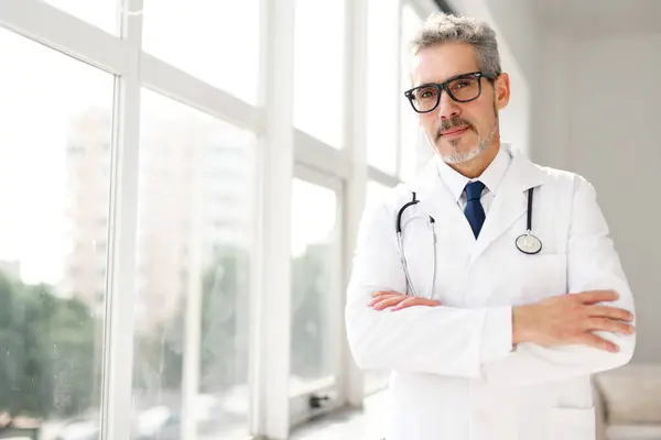 stock image A cheerful grey-haired doctor in a white coat smiles warmly at the camera, his stethoscope visible, representing a friendly healthcare professional ready to serve patients in a modern clinic setting.