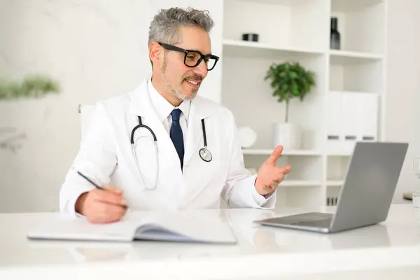 stock image A mature doctor with grey hair is discussing a topic with a patient over a video call, his hand gestures indicating a detailed explanation. Contemporary telemedicine