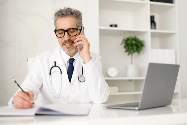 A senior male doctor with grey hair is talking on the phone while writing notes. The setting suggests a modern medical office environment, emphasizing multitasking and patient consultation.