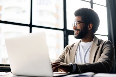 Afro saç stiline sahip neşeli genç bir işadamı arkasında büyük pencerelerden gelen doğal ışıkla yıkanmış bir dizüstü bilgisayarla çalışıyor. Sahne, modern ve dinamik bir çalışma ortamını kapsıyor.