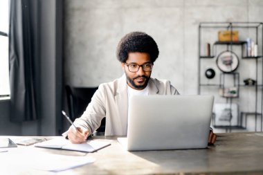 Young Brazilian businessman pensively writes notes, attentively staring at the laptop screen a testament to the meticulous nature of business planning, sitting in modern loft office clipart