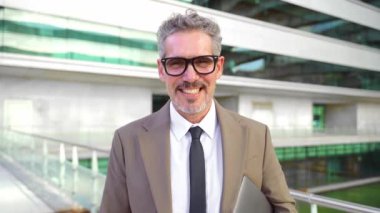 Dışarıda dizüstü bilgisayar tutan kıdemli bir işadamı, rahat tavırları ve modern ofis arkaplanı modern yöneticilerin herhangi bir yerden çalışma esnekliğini gösteriyor.