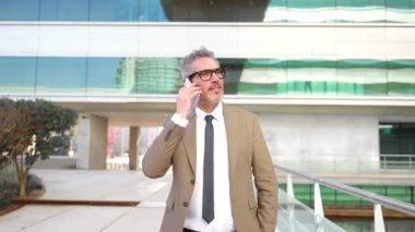 Akıllı iş kıyafetleri içinde kıdemli bir profesyonel hoş bir telefon görüşmesi yapıyor. Yetişkin bir iş adamı arka plandaki modern ofis binasıyla konuşurken yan yana yürüyor ve bakıyor.