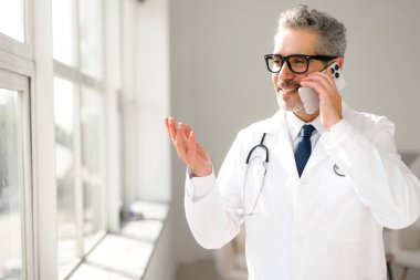 Kendinden emin bir kıdemli doktor telefonda konuşuyor, sanki pencerenin önündeki doğal ışıkla aydınlatılmış karmaşık bir tıbbi sorunu açıklıyormuş gibi el kol hareketi yapıyor, profesyonel bir hasta bakımını uzaktan yönetiyormuş gibi gösteriyor.