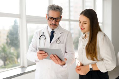Dijital tabletli olgun bir doktor, şehir manzaralı bir pencerenin kenarında genç bir kadın hastayla konuşuyor. Bu, şeffaf ve parlak bir sağlık deneyimini simgeliyor.
