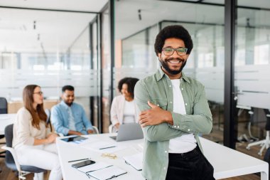 Genç adam modern ofis alanında kollarını kavuşturarak duruyor, rahat ama profesyonel çalışma ortamını sergiliyor, arkasında çeşitli meslektaşlarından oluşan bir ekip verimli ve işbirlikçi bir toplantı yapıyor.