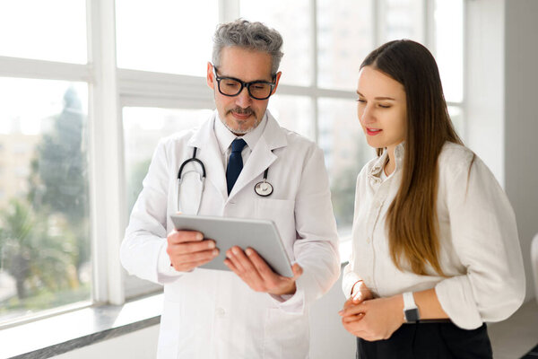 Зрелый врач с цифровым планшетом беседует с молодой пациенткой у окна с видом на город, символизируя прозрачный и яркий опыт здравоохранения