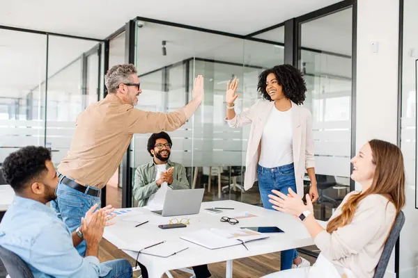 stock image Elation and team spirit are palpable as two colleagues high-five in a contemporary office space, celebrating a collaborative achievement with vibrant enthusiasm. The diverse team applauds happily