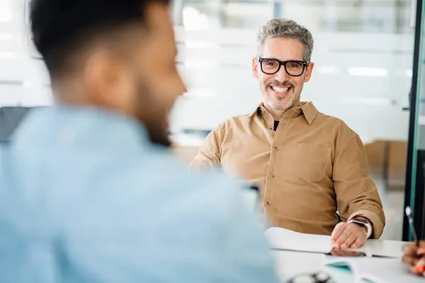 stock image A mature professional man in a tan shirt beams at a colleague out of focus in the foreground, exemplifying a supportive and collaborative office environment during a productive meeting.