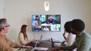 Duvara monte edilmiş bir ekranda gösterilen bir video konferans aracılığıyla uzak meslektaşlarla yüz yüze görüşüp iş birliği yaptıkları bir melez ekip toplantısı. Bağlılık ve esneklik kavramı