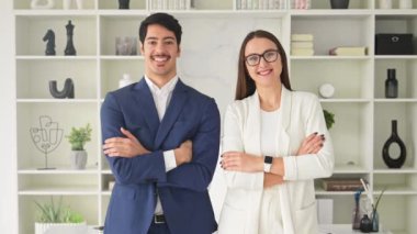 Mavi takım elbiseli bir adam ve beyaz ceketli bir kadın kollarını kavuşturmuş modern ofis ortamında güvenle gülümsüyor. Başarılı bir işbirliği duygusu.