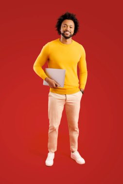 Elinde dizüstü bilgisayarla kameraya bakarken gülümseyen olumlu adamın tam boy görüntüsü. Kırmızı arkaplanda kapalı stüdyo çekimleri