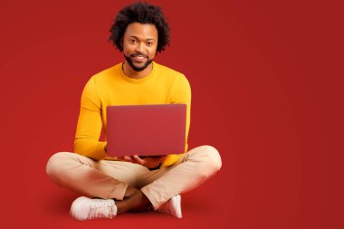 Kırmızı arka planda izole edilmiş dizüstü bilgisayarı tutan ve kullanan mutlu genç sakallı adamın portresi. Erkek ofis çalışanı daktilo ediyor, e-postaları cevaplıyor, oturuyor