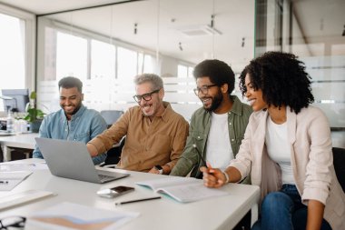 Modern bir ofiste dizüstü bilgisayarla çalışırken çeşitli profesyonellerden oluşan bir grup birlikte güler. Ekip, olumlu takım çalışması ve işbirliği sergileyen bir iş görüşmesine katıldı..