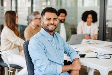 İş toplantısı sırasında modern bir ofiste otururken kameraya gülümseyen kendinden emin genç bir profesyonel. Çağdaş bir iş yerinde takım işbirliği ve verimlilik