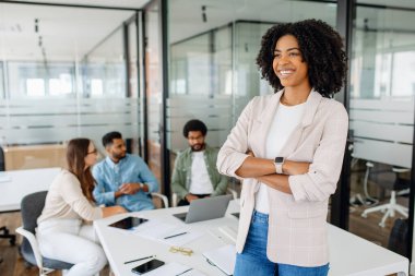 Genç bir bayan profesyonel modern bir ofiste kollarını kavuşturarak gülümsüyor. Arka planda, çeşitli bir ekip bir iş görüşmesi yapıyor, bir proje üzerinde çalışıyorlar..