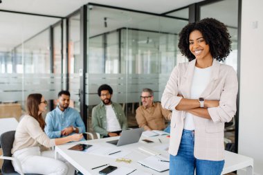 Açık renk ceketli, güler yüzlü bir bayan iş adamı modern bir ofis alanında duruyor. Arkasından, çeşitli bir ekip iş amaçlarına ulaşmak için işbirliği içinde bir tartışmaya girişiyor.
