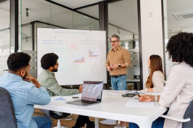 Modern ofiste stratejik bir sunum yöneten işletme yöneticisi. Takım üyeleri aktif olarak dinliyor, şirket planlaması, pazar analizi ve şirket başarısı için yenilikçi karar verme sürecinde yer alıyorlar.