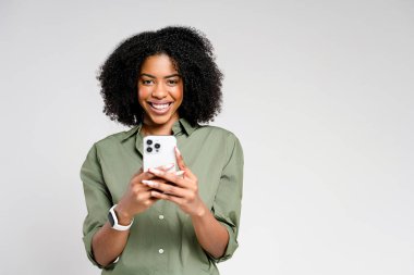 Afro-Amerikan bir kadın akıllı telefonunu kullanırken gülümsüyor, bağlı kalmanın neşesini simgeliyor. Sıradan zeytin gömleği ve akıllı bakışları rahat ama teknolojik bir yaşam tarzını yansıtıyor..