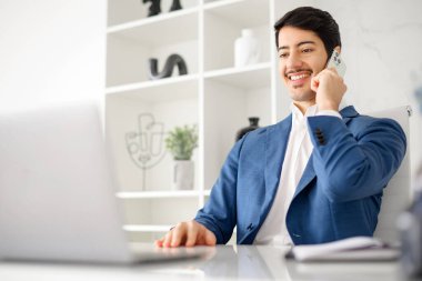 Gülümseyen İspanyol bir işadamı, aynı anda bir telefon görüşmesini yönetirken, aynı zamanda parlak ve çağdaş bir ofis alanında,